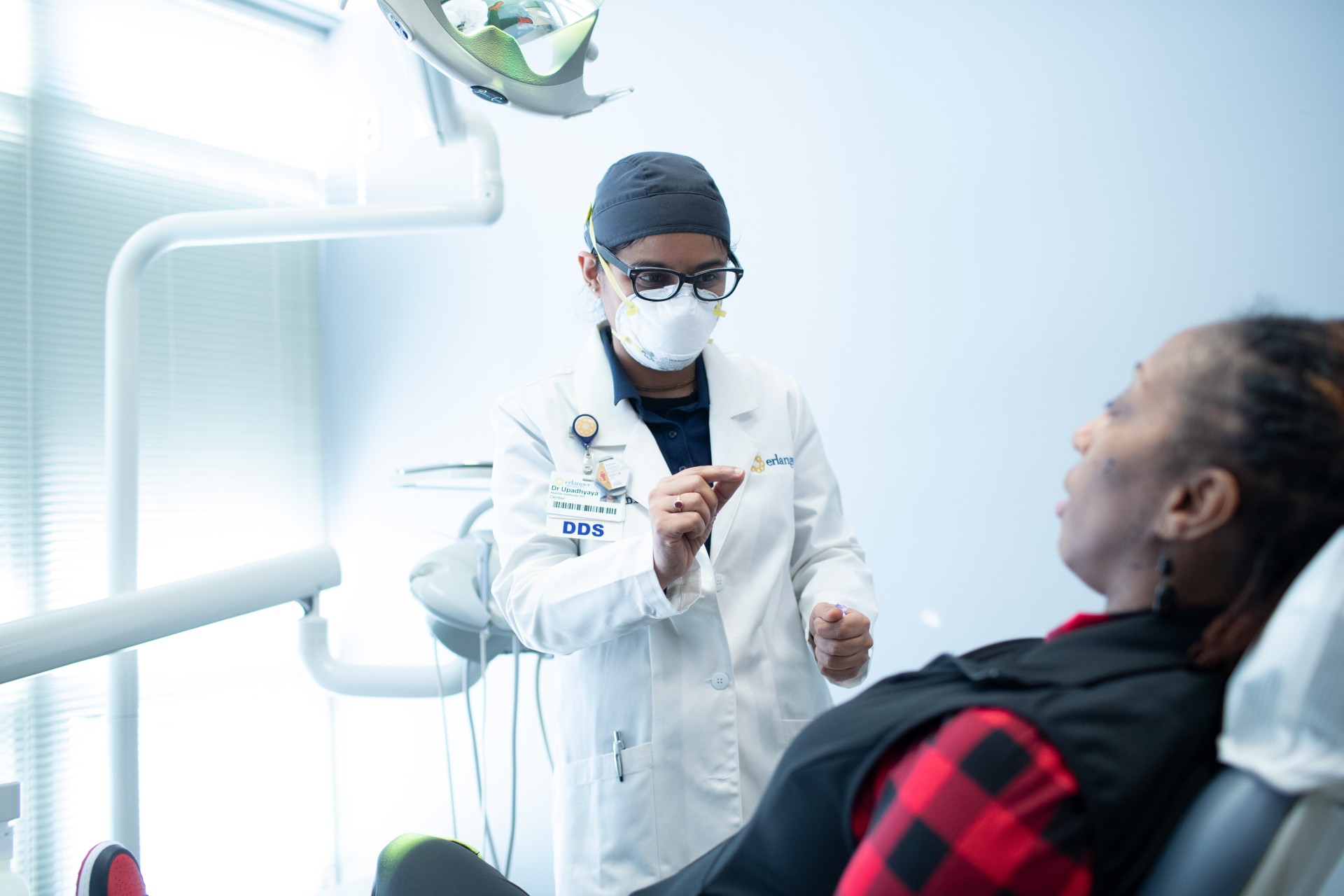A dentist wearing a white lab coat, N95 mask, and glasses consults with a patient seated in a dental chair in a bright, modern exam room