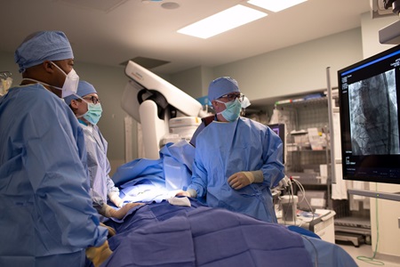A surgical team in blue scrubs and masks performs a procedure in a high-tech operating room while monitoring a digital screen displaying a medical imaging scan of a heart