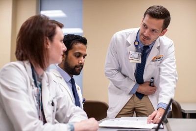 Three medical professionals in white lab coats collaborate around a desk, with one doctor leaning over to point at a document in a binder while discussing it with two seated colleagues
